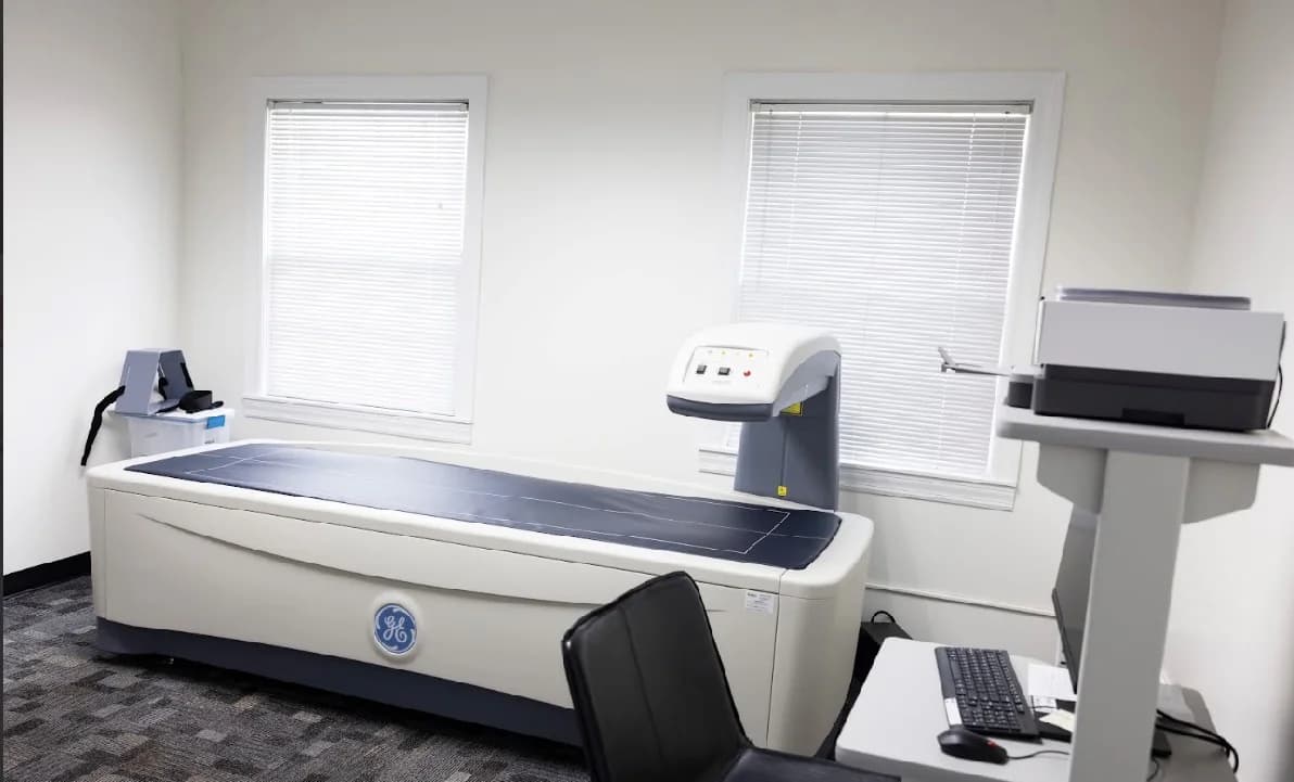 A bright room with a DEXA body composition scanner, a computer workstation and two windows with white blinds.
