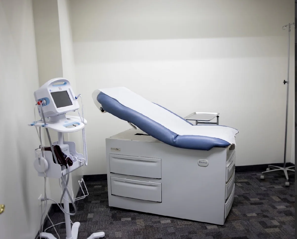 A bright exam room with a padded exam table, a vital-signs monitor on a stand and an IV pole.